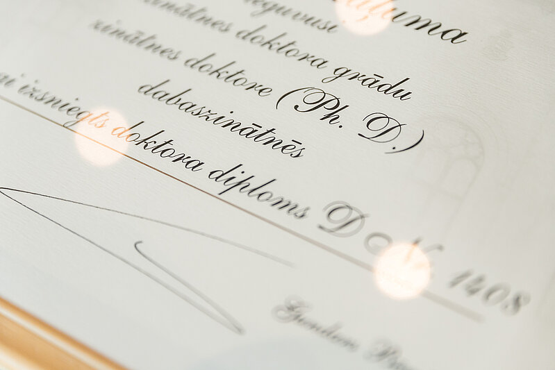 Tuvplāns doktora diploma ar kaligrāfisku tekstu latviešu valodā, kurā redzami vārdi "doktora grādu", "Ph.D.", "dabaszinātnēs" un "doktora diploms".