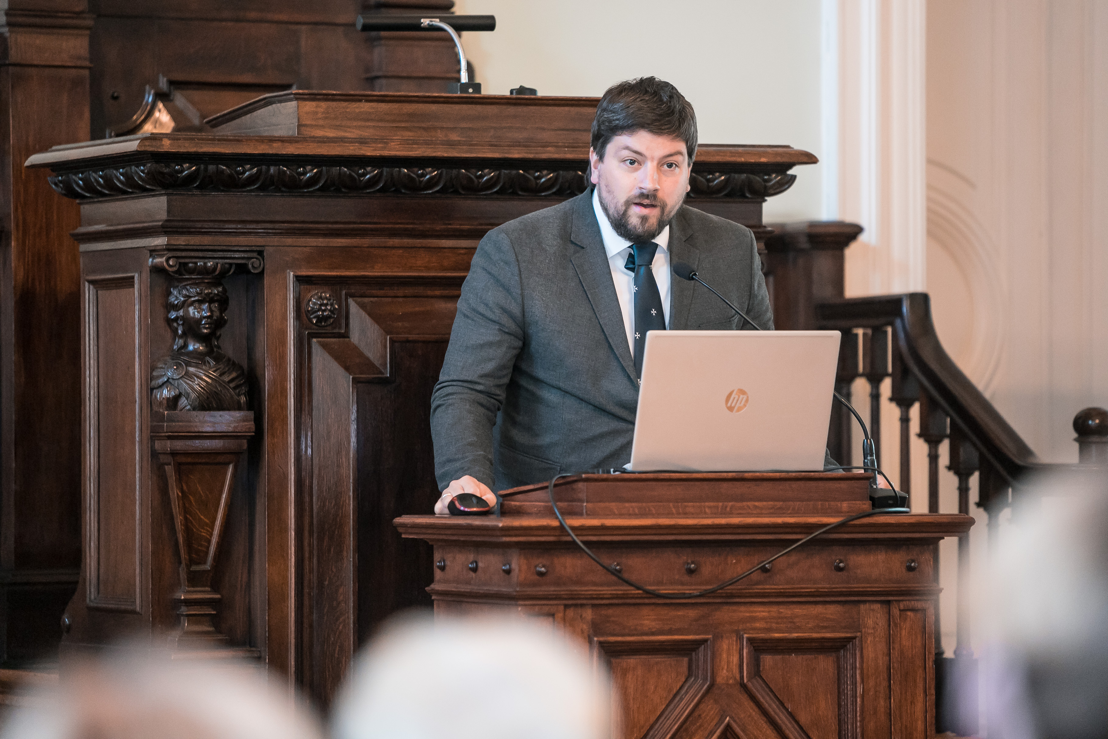 LU Zinātņu prorektors uzstājas ar prezentāciju pie tribīnes, izmantojot klēpjdatoru un mikrofonu auditorijas priekšā.
