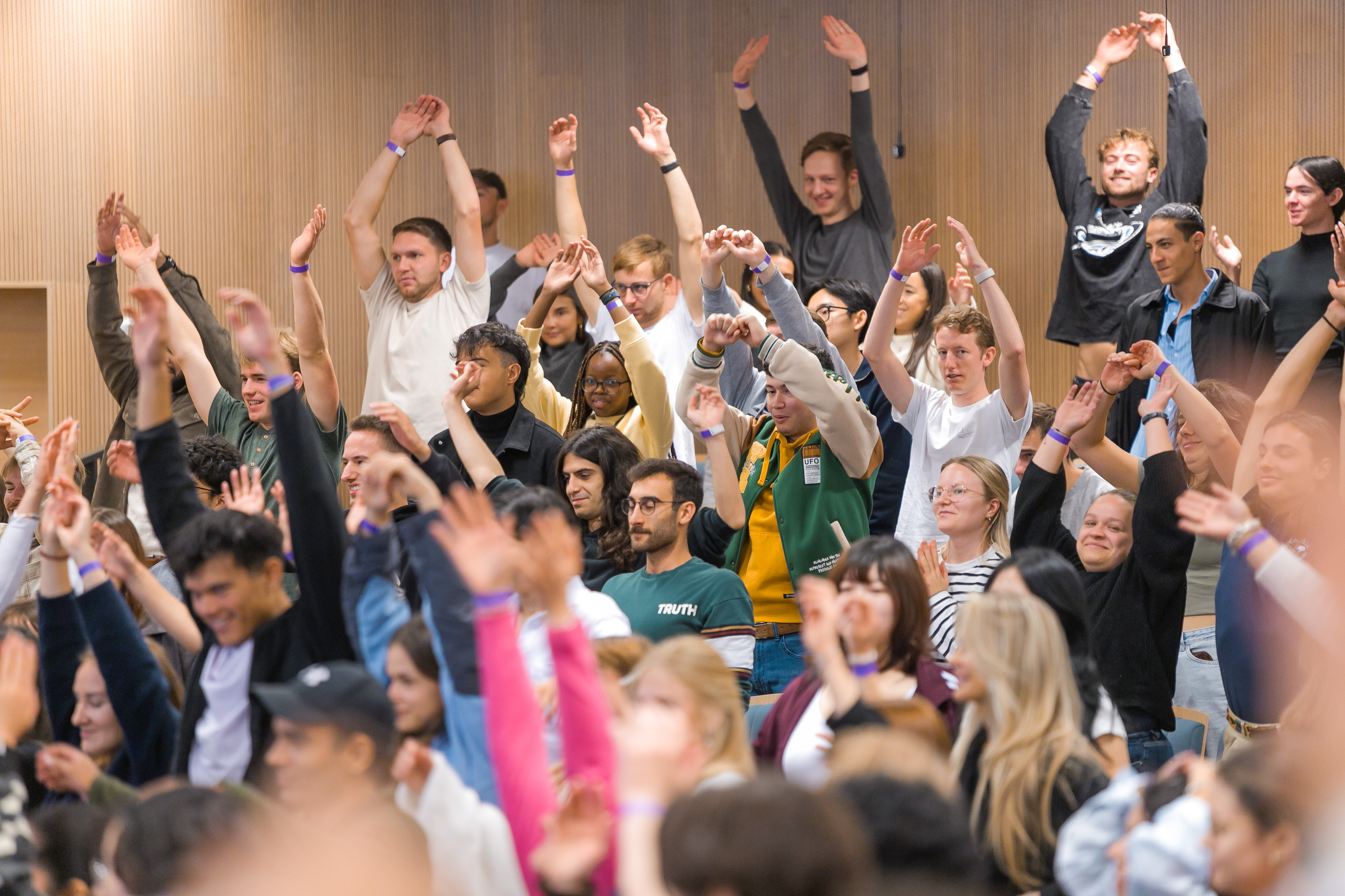 Liela cilvēku grupa auditorijā paceļ rokas, aktīvi piedaloties pasākumā