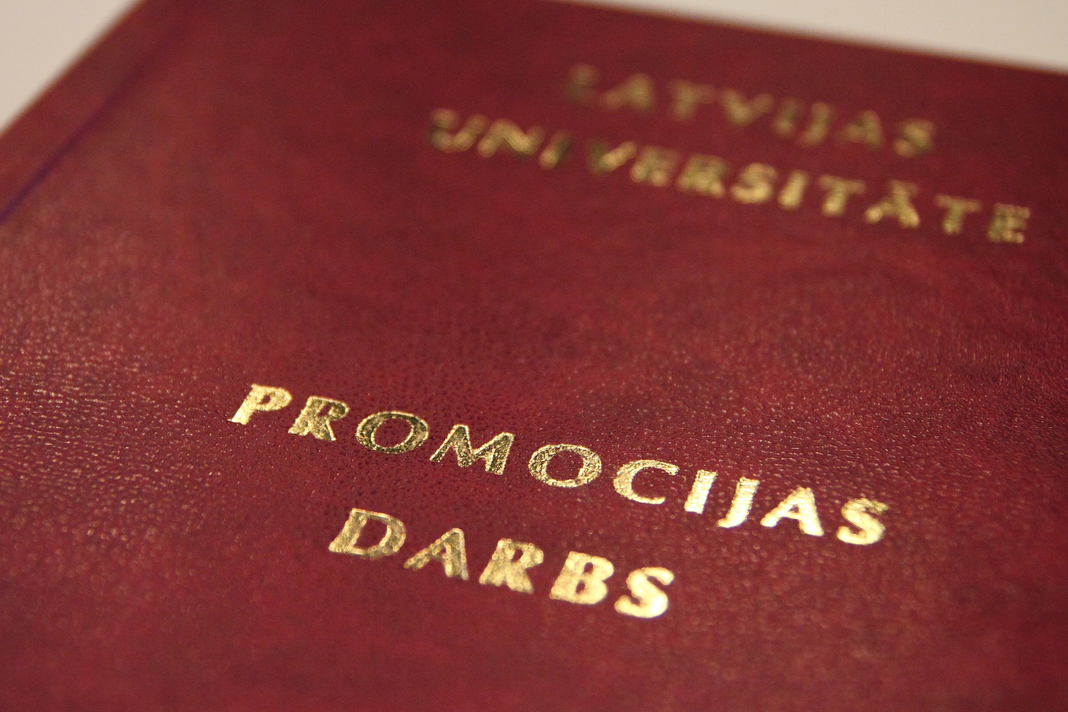 Latvijas Universitātes promocijas darbs tumši sarkanā vākā ar zelta burtiem, novietots uz galda blakus atvērtai grāmatai. Fonā redzami grāmatu plaukti.