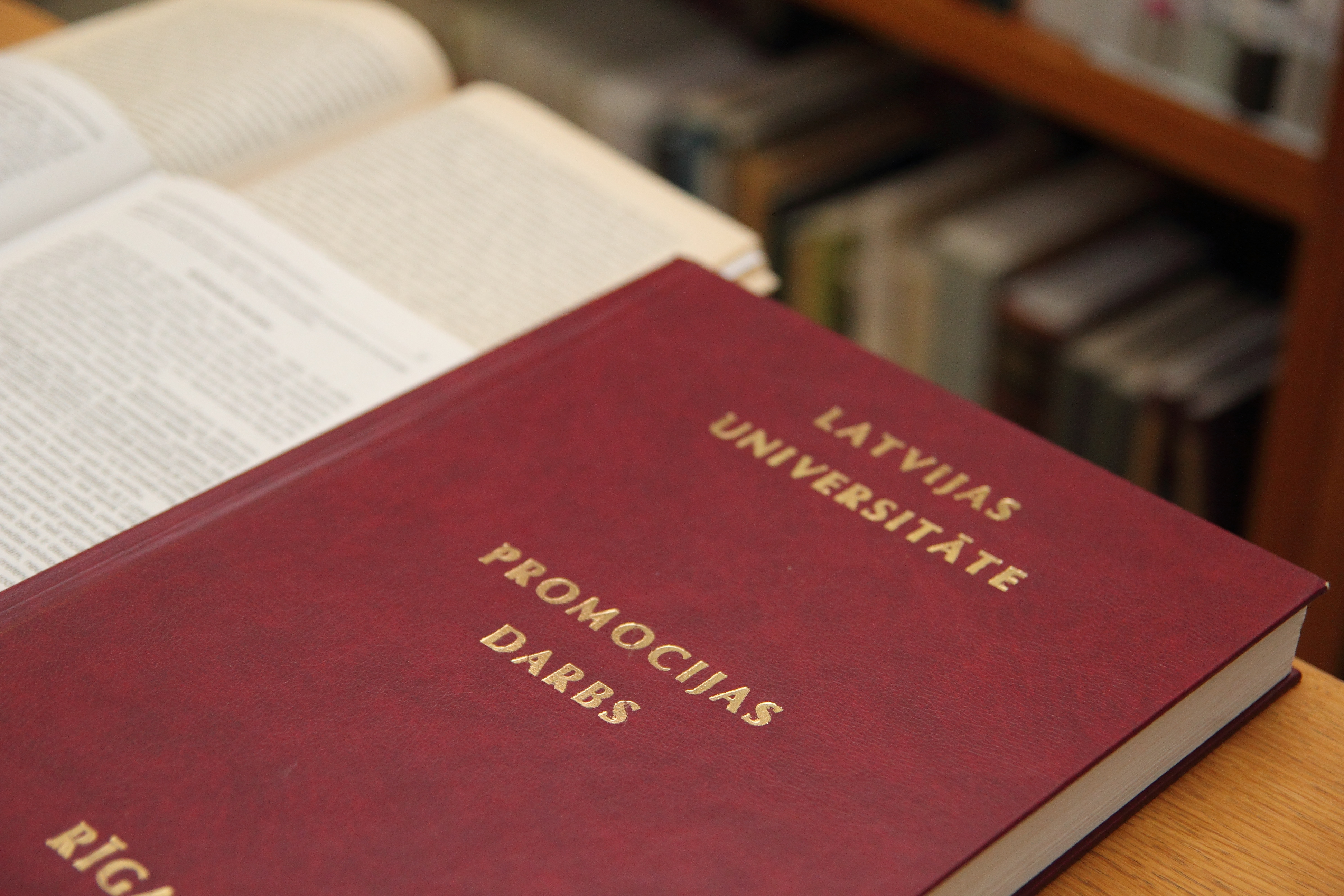Attēlā redzama sarkana cietā vāka grāmata, kas novietota uz atvērtām lapām. Uz vāka ar zelta burtiem ir rakstīts “LATVIJAS UNIVERSITĀTE” un “PROMOCIJAS DARBS”, kas norāda, ka tā ir doktora disertācija no Latvijas Universitātes. Grāmata izskatās oficiāli un profesionāli iesieta.  Fonā redzami izplūduši grāmatu plaukti, kas rada bibliotēkas vai akadēmiskas vides iespaidu. Kopumā attēls atspoguļo augstākās izglītības un zinātniskās pētniecības kontekstu.