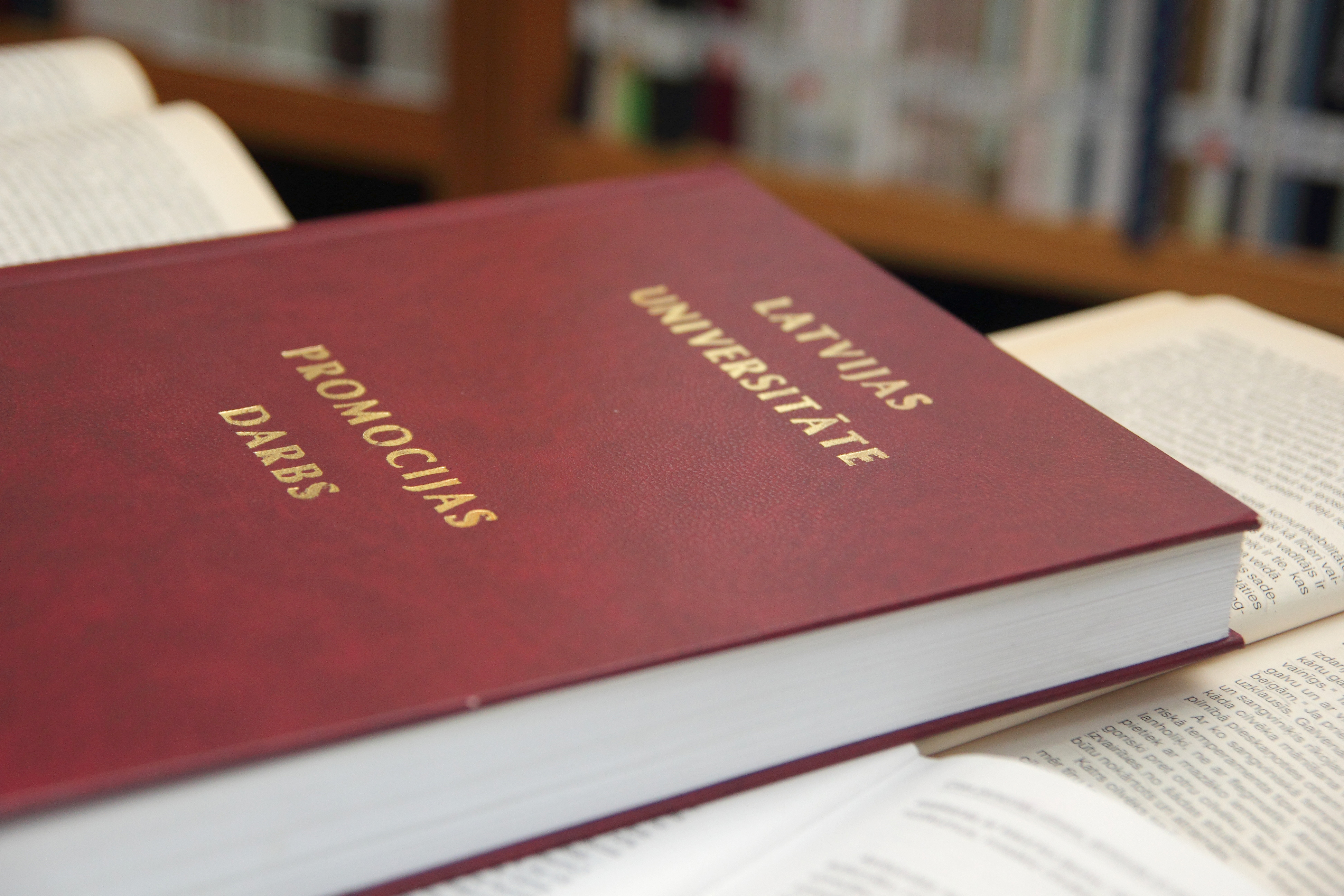 Latvijas Universitātes promocijas darbs tumši sarkanā vākā ar zelta burtiem, novietots uz galda blakus atvērtai grāmatai. Fonā redzami grāmatu plaukti