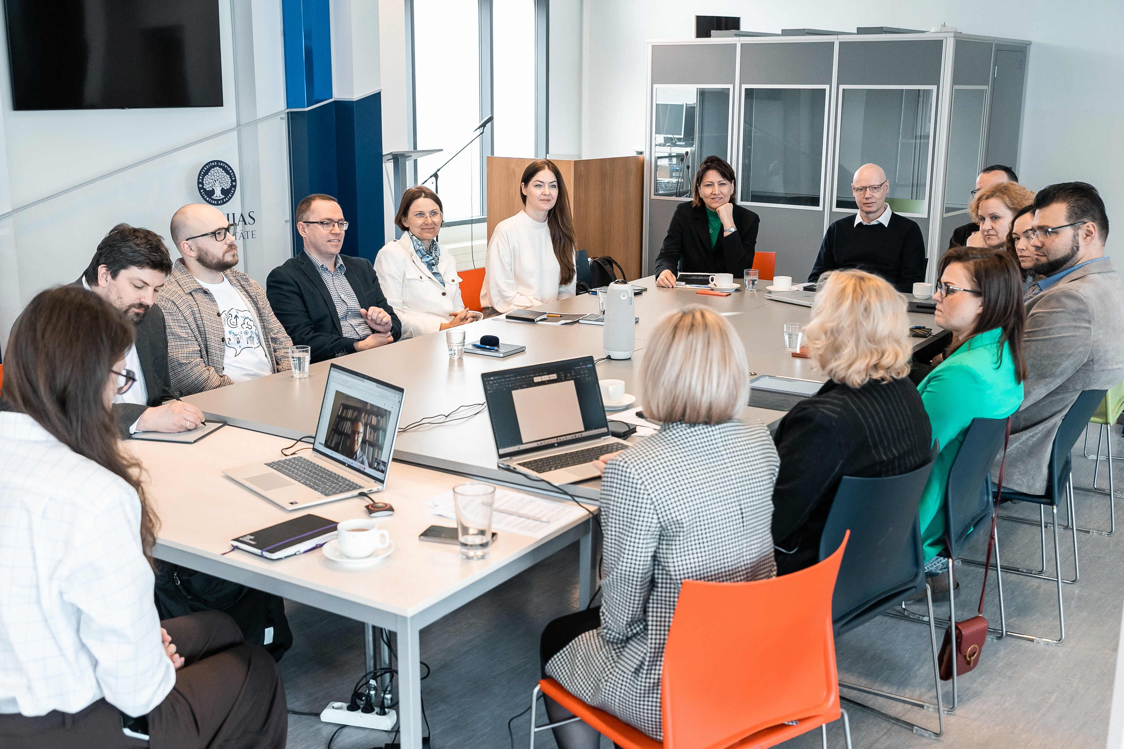 A group of people sitting around a conference table in a modern office space and participating in a meeting; laptops, documents and coffee cups are visible on the table