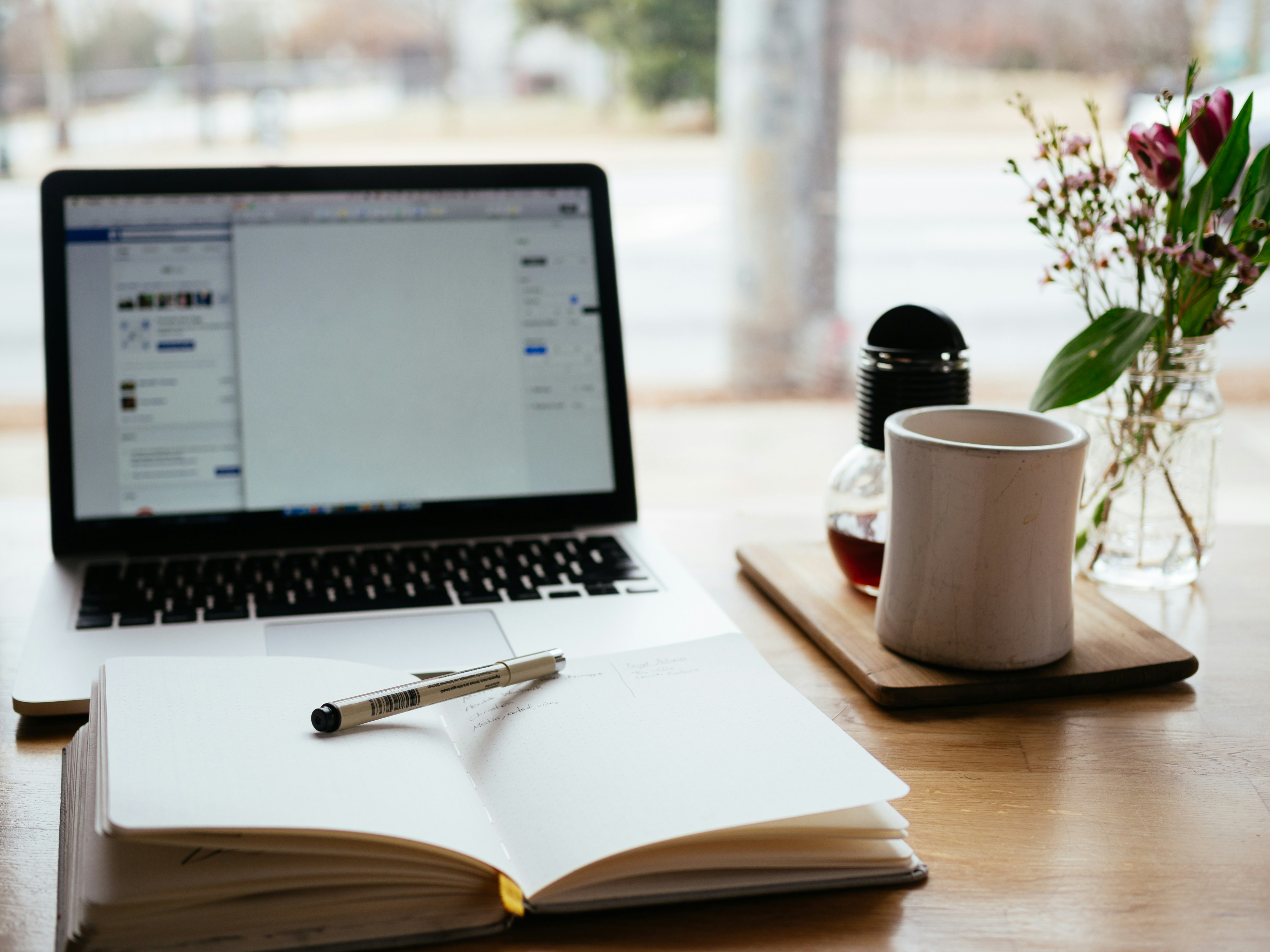 An open laptop sits on a desk next to a notebook with a pen. On a wooden tray, there is a mug, a small bottle, and flowers, creating the impression of a workspace or study environment