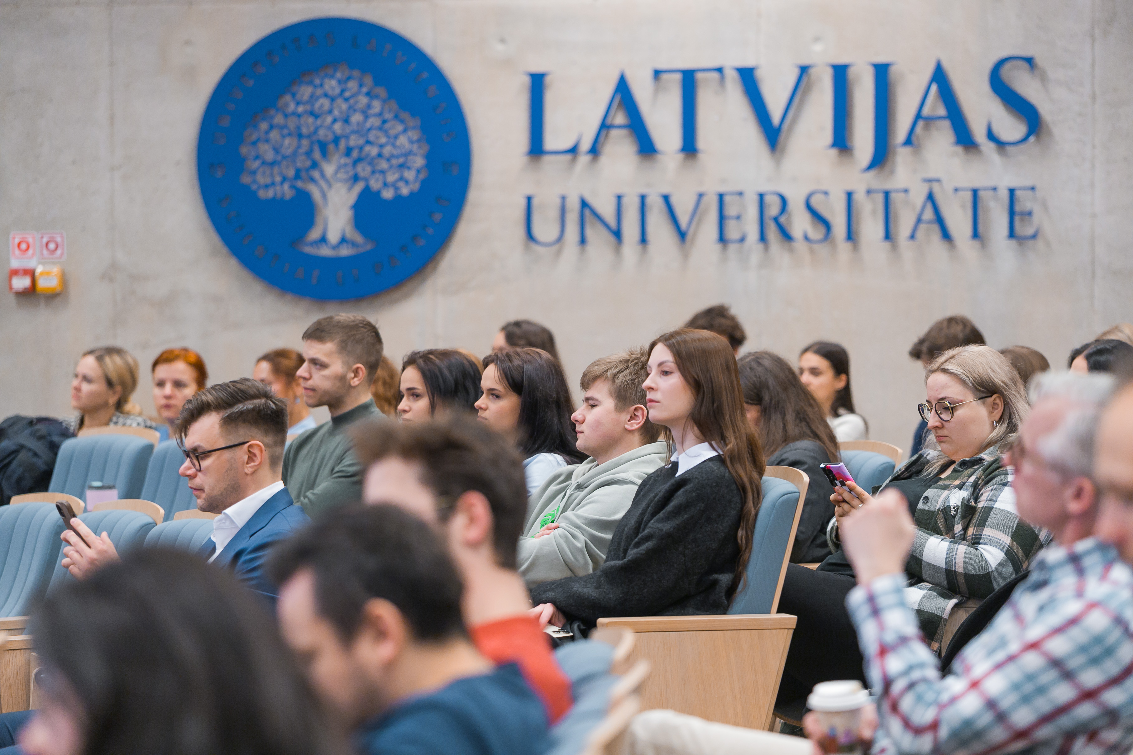 Auditorija ar studentiem un apmeklētājiem, kas sēž krēslos un klausās pasākumu Latvijas Universitātē. Fonā redzams universitātes logotips un nosaukums "Latvijas Universitāte"