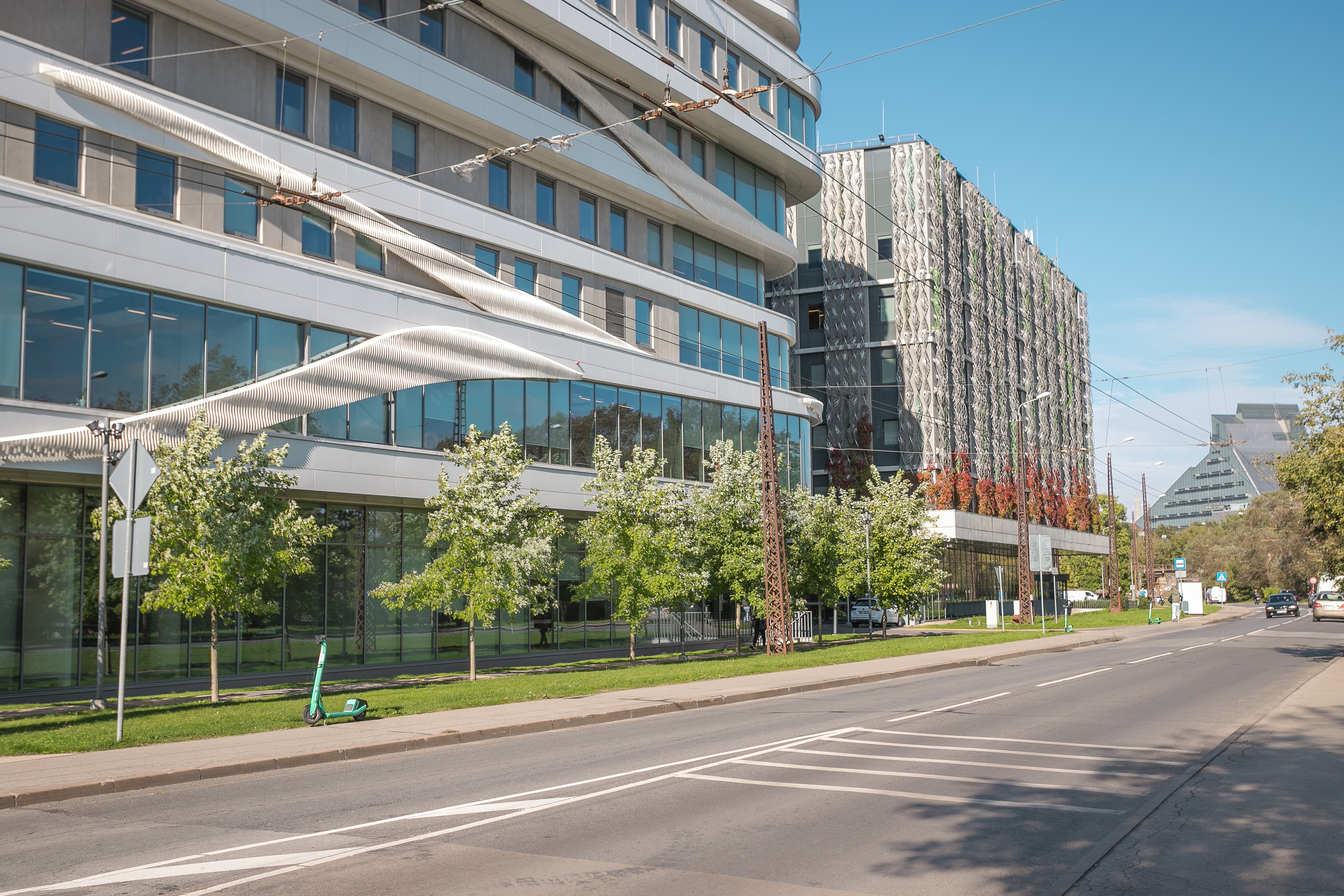 Plata pilsētas iela ar mazu satiksmi. Kreisajā pusē redzama moderna stikla un betona biroju ēka ar nojumi virs ietvja, fonā augstāka daudzstāvu ēka. Ietvi rotā zaļi koki. Fotogrāfija uzņemta saulainā dienā ar skaidru zilu debesu fonu