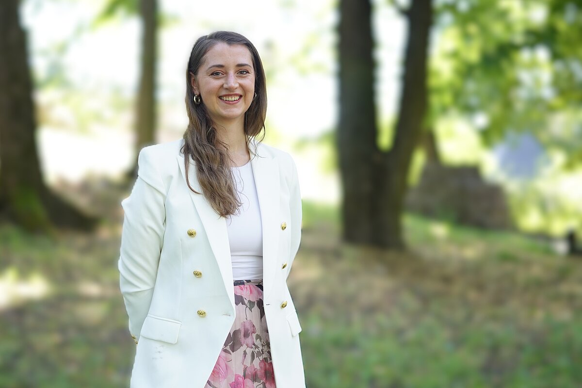 Laura Ansone baltā žaketē ar zelta pogām un rozā ziedu svārkos, stāv parkā zaļu koku fonā.