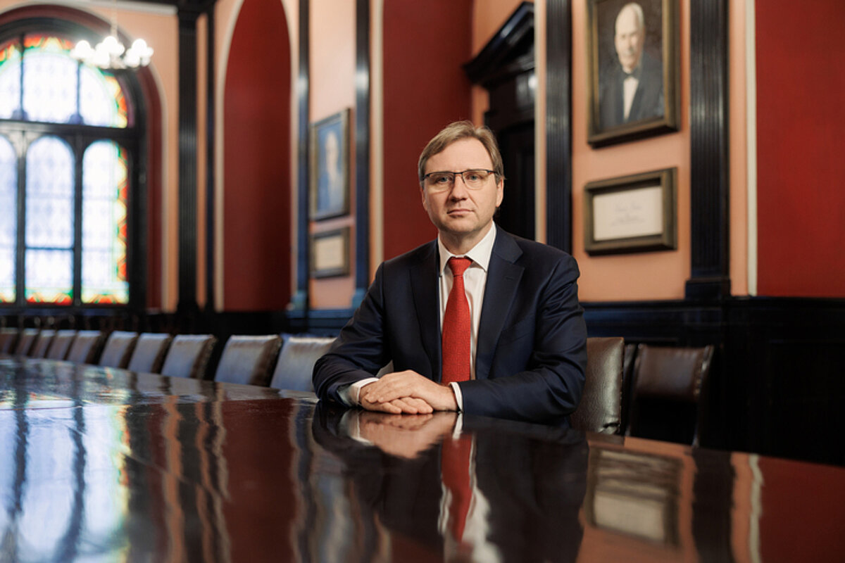 Latvijas Universitātes reftors prof. Gundars Bērziņš ar brillēm tumšā uzvalkā un sarkanā kaklasaitē sēž pie liela koka konferences galda vēsturiskā zālē ar vitrāžu logiem un portretglazniecību sienās.