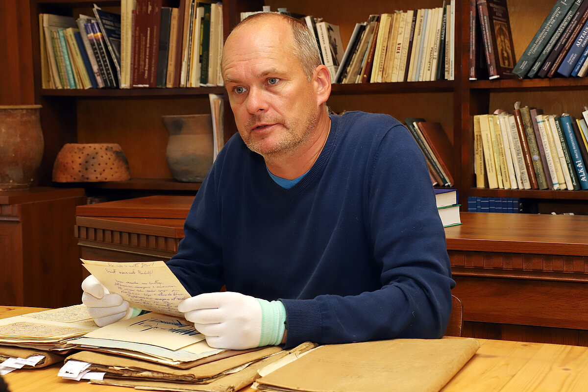 Attēlā redzams vīrietis, kurš sēž pie koka galda, iespējams, bibliotēkā vai pētniecības telpā. Viņš ir ģērbies tumši zilā džemperī pār gaiši zilu kreklu un valkā baltas cimdus, rūpīgi turot rokās rokrakstītu dokumentu vai vēstuli. Uz galda priekšā ir vairākas papīru un mapju kaudzes. Aizmugurē redzama koka grāmatu plaukts ar daudzām grāmatām un dažiem keramikas priekšmetiem, kas liecina par akadēmisku vai arhīva vidi