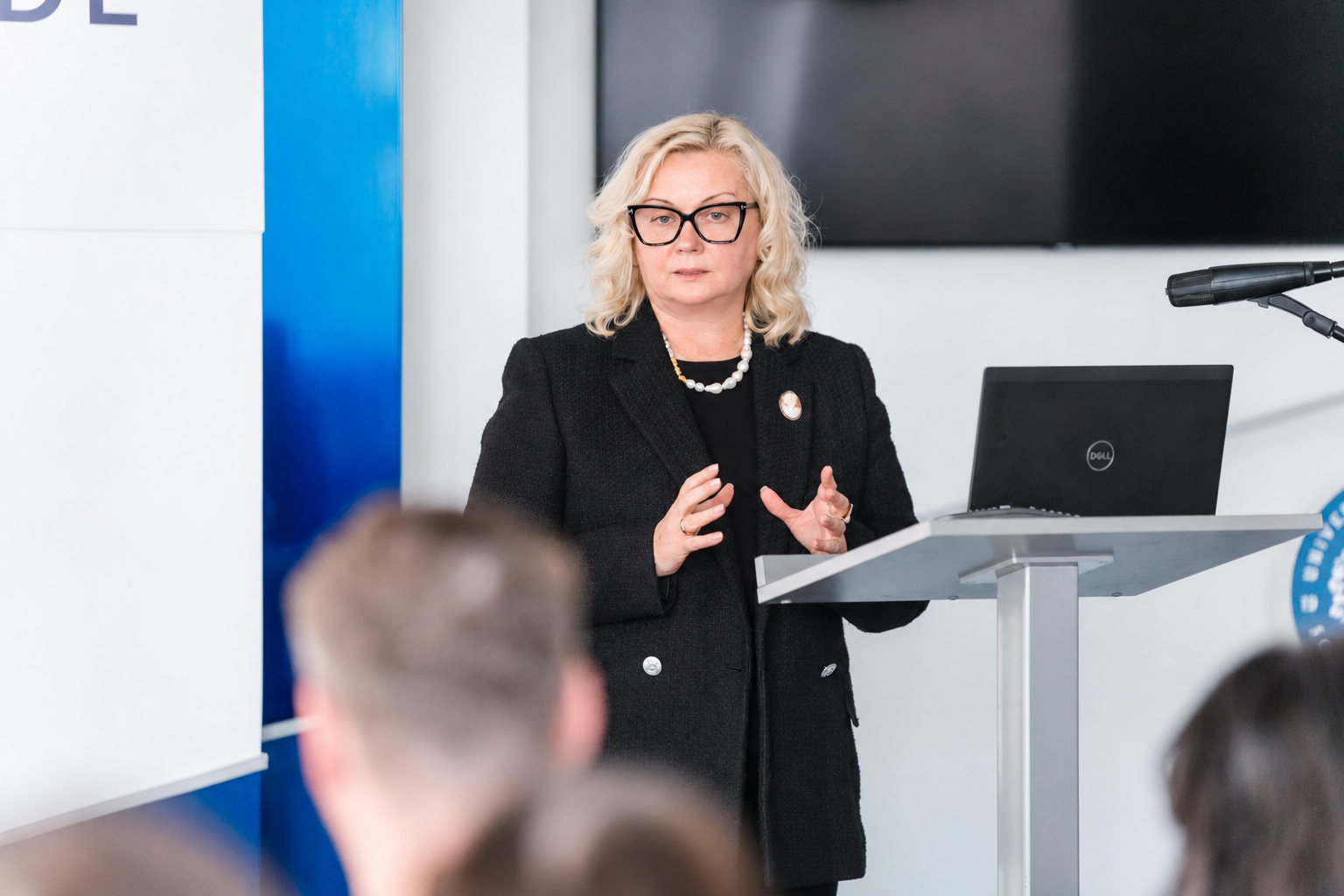 LU Doktorantūras skolas direktore prof. Zanda Rubene stāv pie tribīnes un uzstājas konferencē. Uz tribīnes atrodas klēpjdators un mikrofons, priekšplānā redzami klausītāji.